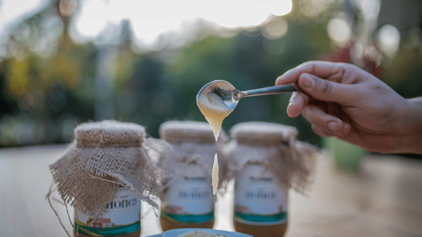 A close up shot of 3 sealed jars of honey with a bowl of honey being poured by a person - Karma Lakelands.