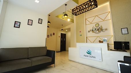 A reception area with a dark sofa and a white counter featuring a logo, illuminated by modern lighting fixtures - Asian Suites Cyber Park, Gurgaon