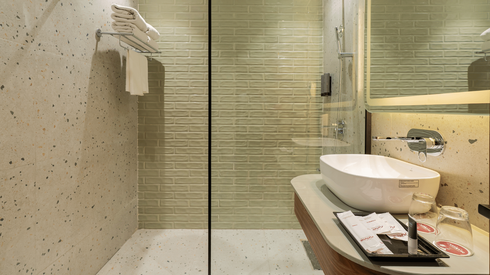 A bathroom with a walk-in shower behind a glass partition, a big mirror with a wash basin, toiletries on a counter top, and fresh towels on a wall shelf | Summit by the Ganges Beach Resort & Spa, Rishikesh