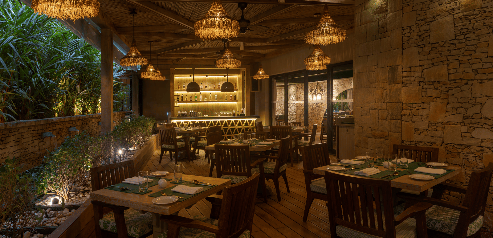 A dimly-lit dining area at the Mila Restaurant with lush green trees on one side at The Evren, Vagator.