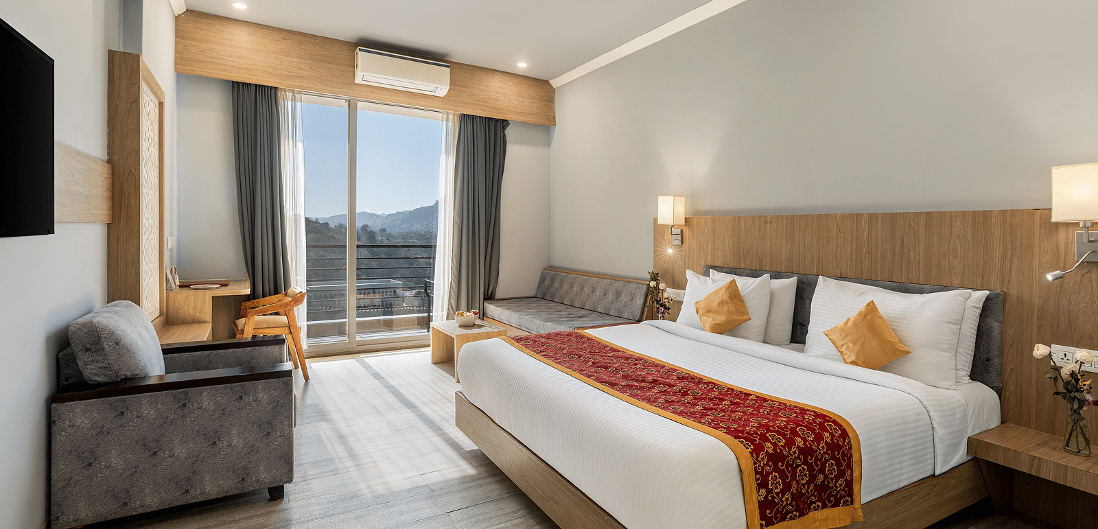A hotel room with a bed, seating area, desk, and view through a large window - Via Lakhela Resort & Spa, Kumbhalgarh