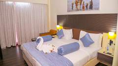 A hotel guestroom at Ambar Sarovar Portico, Gandhidham is furnished with a king size bed, bedside lamps, and a decorative painting above the headboard.