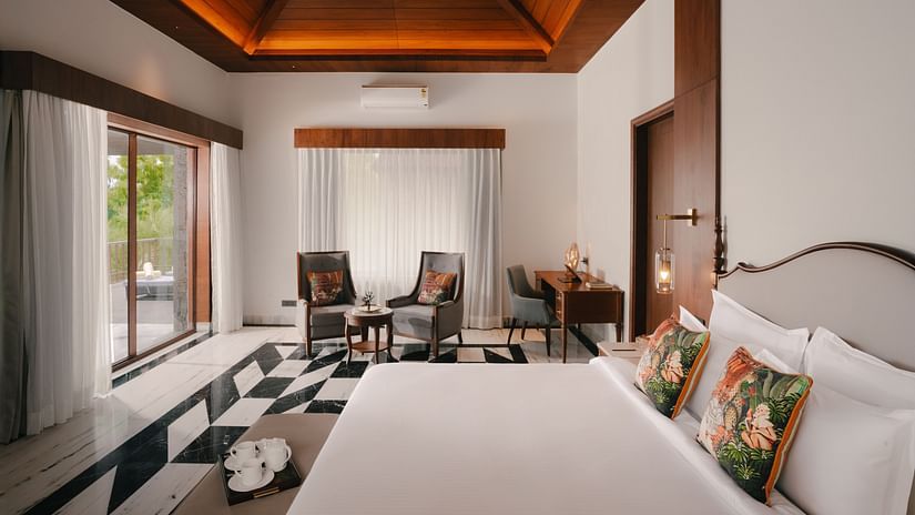 A hotel room with a bed featuring patterned cushions, a seating area with chairs, and a view of the outdoors through a window.