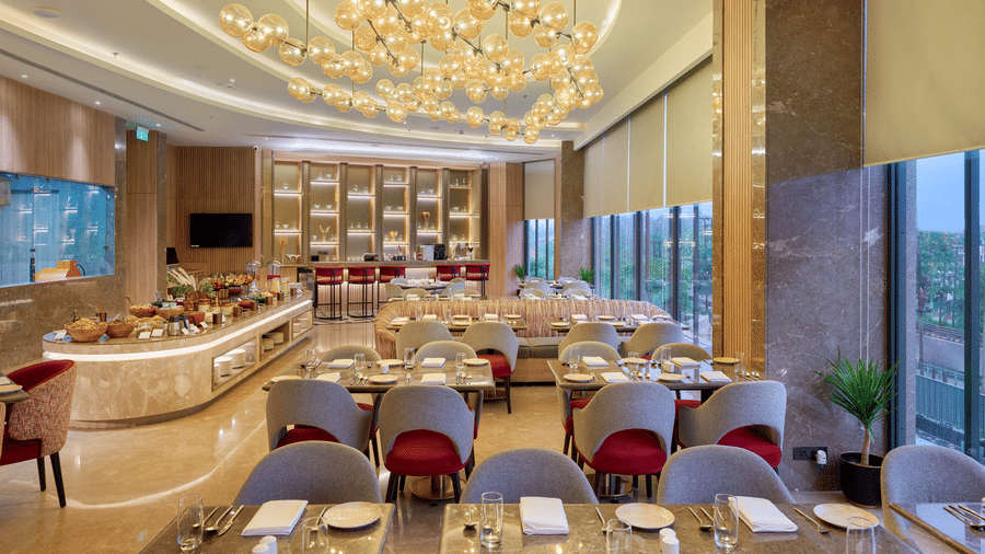 A restaurant interior at KPG Sarovar Portico, Raj Nagar Extension, Ghaziabad featuring dining tables, chairs, a decorative ceiling fixture, and large windows.