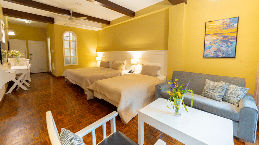 A spacious family suite featuring multiple beds, warm yellow walls, and exposed dark wood ceiling beams - Johnson Lodge & Spa, Manali