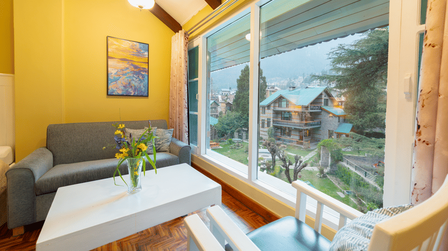 An indoor seating area with a white coffee table and grey sofa, offering a clear view of the lodge's lush surroundings - Johnson Lodge & Spa, Manali
