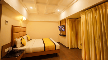 Guest room at Hotel Grand Sabarees Madurai featuring a double bed with yellow runner, wall-mounted TV, bedside tables, and yellow window drapes.