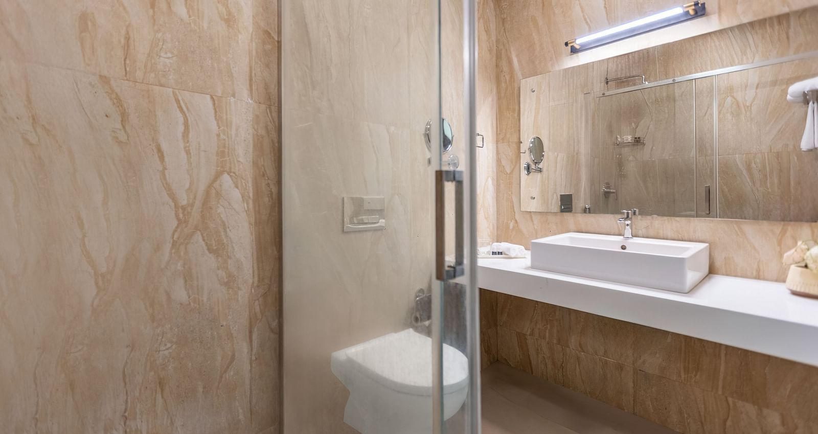 A bathroom with beige and brown textured walls, a glass shower stall, a white rectangular sink, and a mirror - Mango Hill Resort, Coimbatore