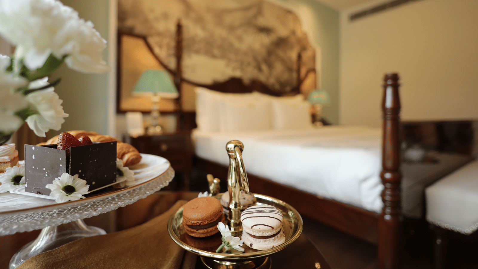A blurred view of a bedroom with a four-poster bed and a mural, with a close-up of a tray with pastries and a small cake on a table in the foreground | Dileep Kothi, Jaipur