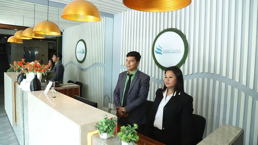 Two individuals stand behind a reception desk in a well-lit area with decorative lighting - Asian Suites IFFCO Metro, Gurgaon