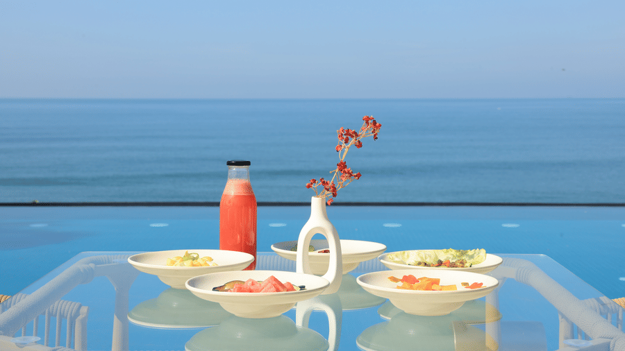 A table overlooking an infinity pool and the ocean, set with a bottle, flower vase, and bowls of food at hotel SUKAVI.