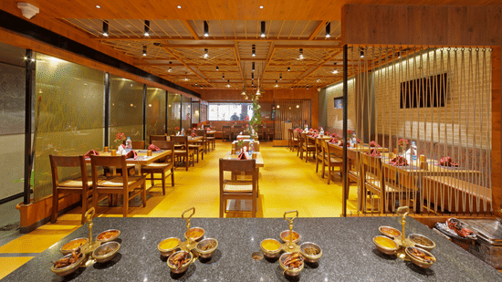 A wide angle view of spacious and inviting restaurant at Icon Boutique Hotel by Bhagini, Domlur, with wooden tables and chairs, a wooden ceiling, and a buffet spread.