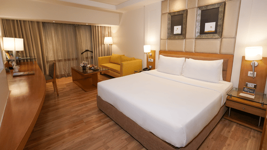 A room at Park Plaza Ludhiana featuring a king bed with white bedding wooden furniture a yellow sofa warm lighting and a work desk area in a modern setting.