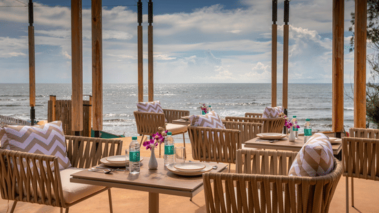 A well arranged tables and chairs at In-House Restaurant with sea waves crashing on the beach in the background