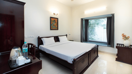 A room in Sanctum Suites Indiranagar, Bangalore, showcasing a double bed with white bedding, a bedside table with a lamp, an electric kettle placed on a table, a chair, and a window providing natural light.