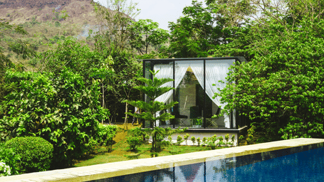 A contemporary glass house pavilion with white drapes set in a lush green garden beside a swimming pool, with rocky hills in the background.
