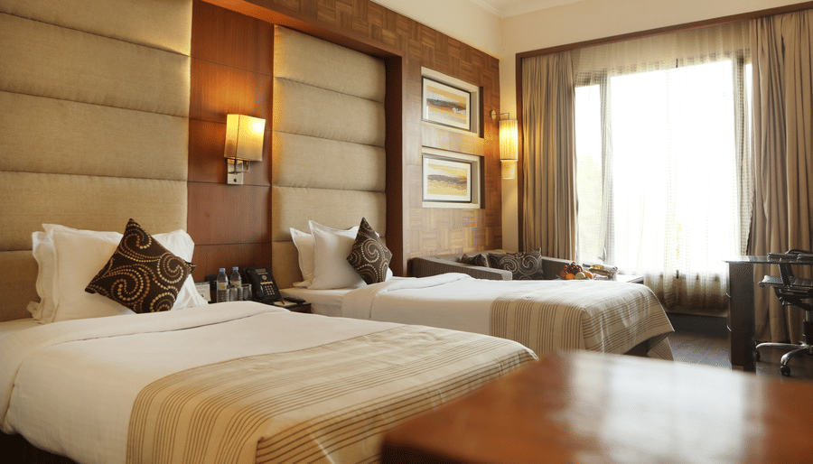 Deluxe Rooms at The Bristol Hotel Gurgaon, Rooms in Gurgaon, featuring twin beds, spreads, pillows, tables, and a window coveed with curtains.