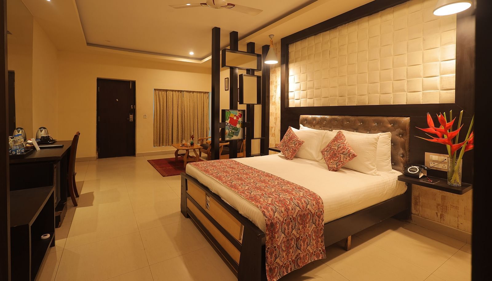 An image of a bedroom with bedside table, a set of table and chairs, flat TV screen, and AC- Sathyam Grand Resort