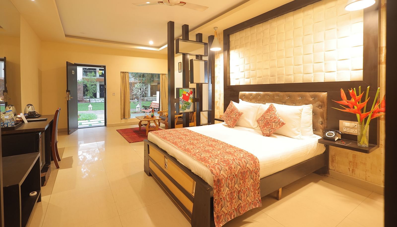 An image of a bedroom with bedside table, a set of table and chairs, flat TV screen, AC and open door- Sathyam Grand Resort