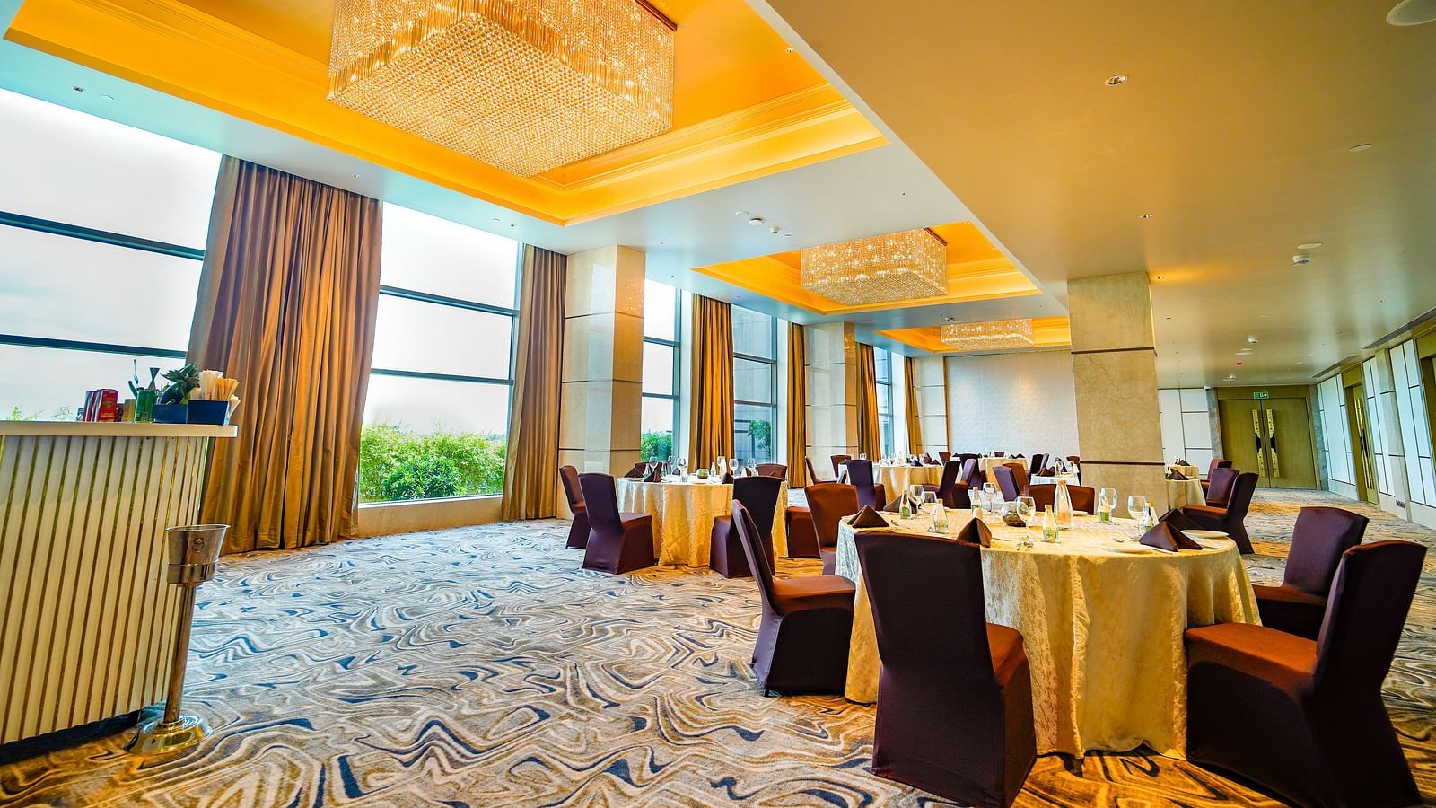 A slightly lower angle shot of a  banquet hall with round tables, elegant chandeliers, and natural light from large windows - Grand Madurai by GRT Hotels
