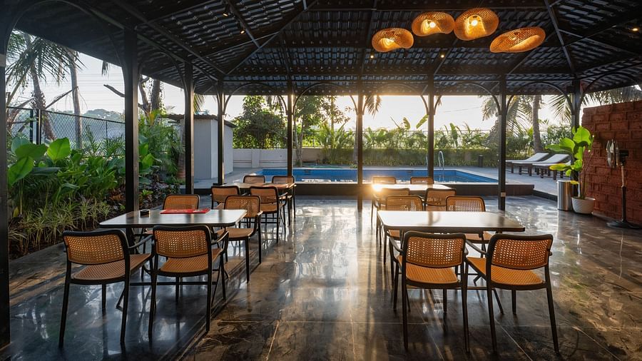 An outdoor dining area under a dark, peaked roof, open to the surrounding scenery. Several wooden tables and chairs are neatly arranged, with a glimpse of a swimming pool visible in the background under the bright sky at Kssemya
