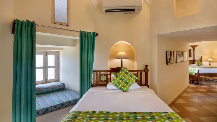 A room with a single bed, a window seat, a framed artwork, and an arched doorway leading to another room in Tijara Fort-Palace - 19th Century, Alwar.