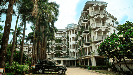 Exterior view of Hotel Sonar Bangla, Tarapith, featuring multi-storey architecture, tall palm trees, parked vehicles, and landscaped surroundings.
