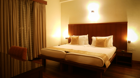 Double bed in the One-Bedroom Suite at Vedic Village Spa Resort, featuring a wooden headrest, warm lighting, wooden flooring, work desk, and closed drapes over a large window.