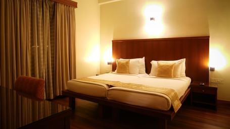 Double bed in the One-Bedroom Suite at Vedic Village Spa Resort, featuring a wooden headrest, warm lighting, wooden flooring, work desk, and closed drapes over a large window.