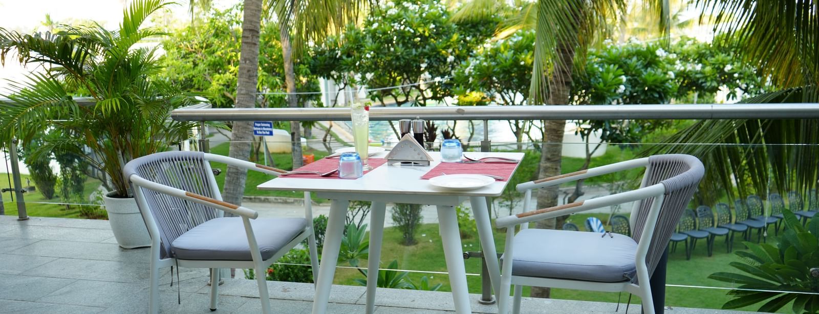 An outdoor dining table set with plates, cutlery and glasses beside greenery.