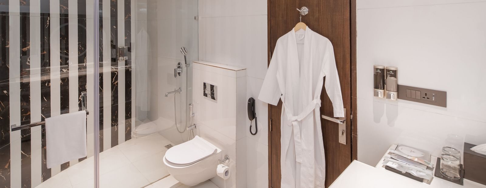 An image of a bathroom with bathrobe hanged on a door, mirror on the wall, cabinet filled with towels and toiletries, and a commode - Brij Eternity, Vrindavan