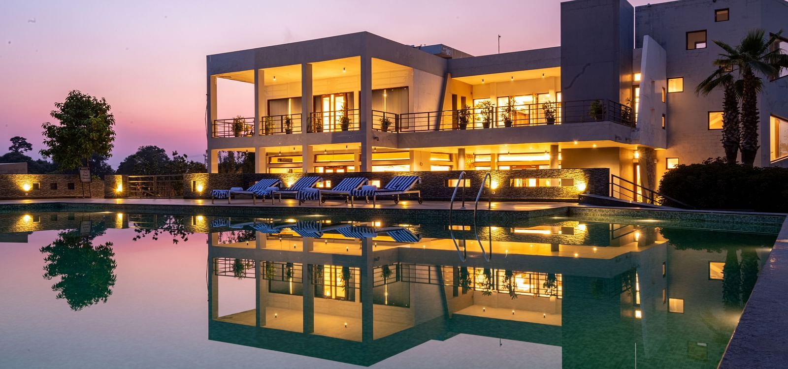 Facade view of DLS Forest Vines Resort, with warm light glowing from its windows and doors, reflected on the shimmering swimming pool beneath a clear sky.