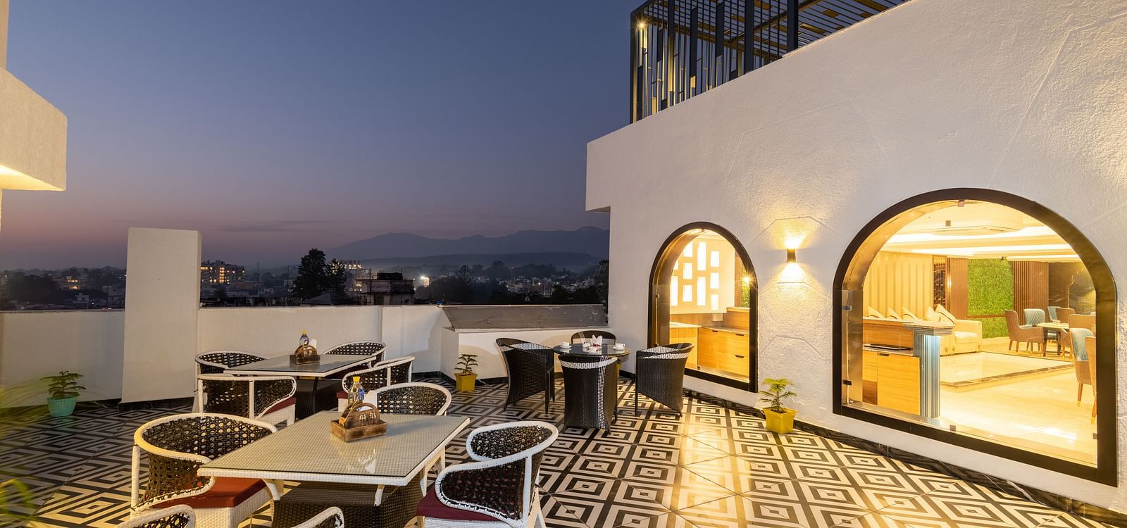 Image of the rooftop restaurant at DLS Grand Luxotica, Dehradun during evening featuring chairs and tables arranged on a well-furnished floor