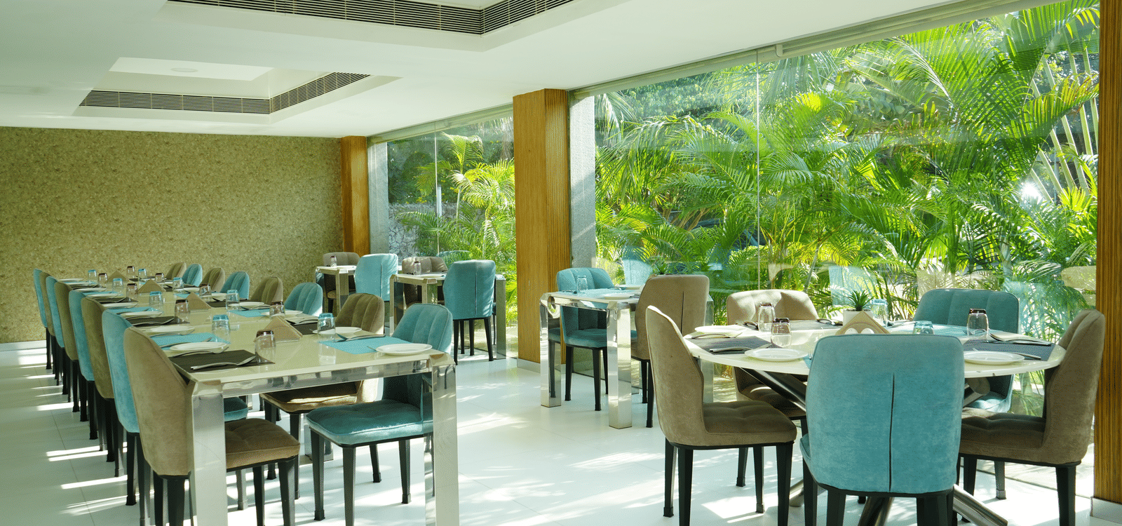 The seating area in L’attitude 49 restaurant in Mamallapuram featuring turquoise coloured chairs, round and long tables, in front of window with lush green views - Grande Bay Resort & Spa, Mamallapuram