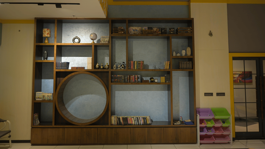 A wooden display shelf in the indoor games area showcasing percussion instruments, board games, and decorative items at Hotel Sonar Bangla Mayapur.