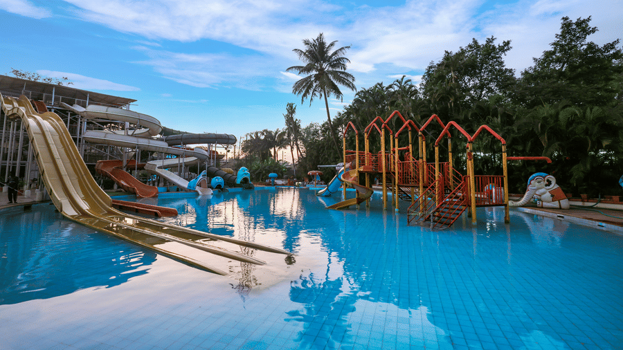 Outdoor water park swimming pool with large slides, water play structures, and tropical palm trees visible under a bright sky at Kumar Resort by Turtle.