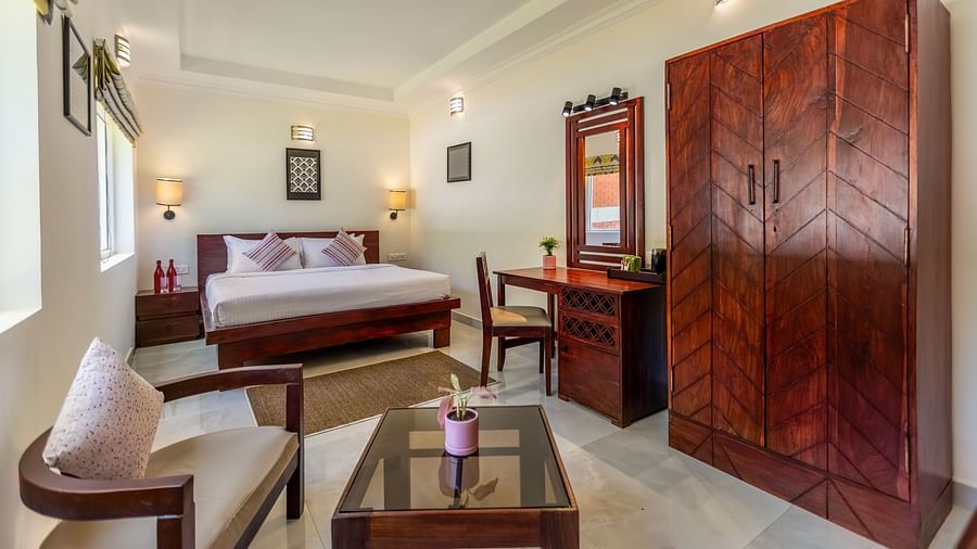 A bright, clean hotel room with a bed, a wooden desk and chair, a large wooden wardrobe, and a seating area with a glass-topped coffee table - Mango Hill Resort, Coimbatore