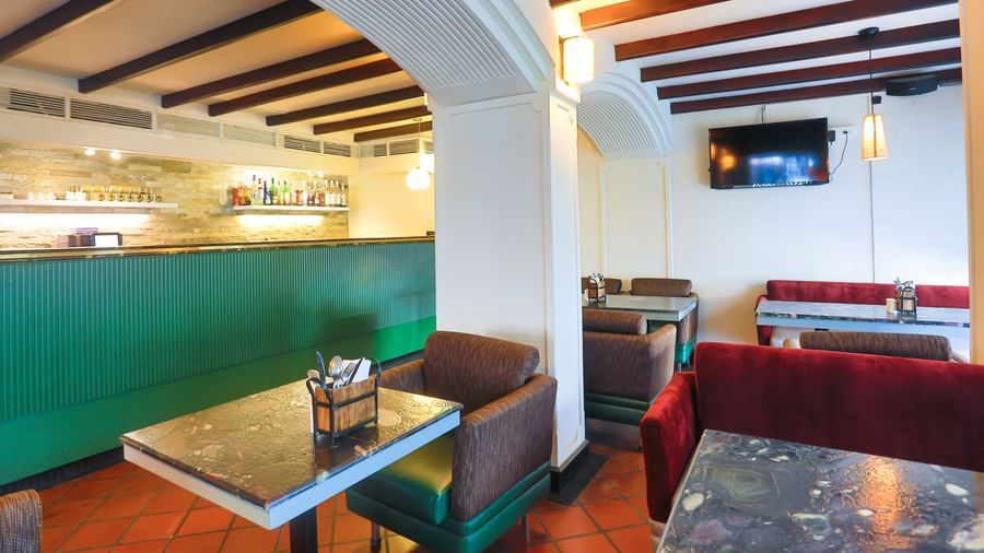 A cafe interior at Shilton International, Kochi, with tables, chairs, and booths, featuring a counter with shelves and a television.