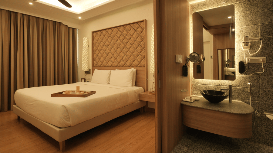 A spacious bedroom with a king size bed, upholstered headboard, wooden flooring, and an attached open bathroom area with a glass partition in the Presidential Suite at Clarks Amer, Jaipur.