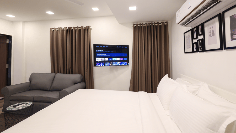 A spacious bedroom featuring a neatly-made bed, a wall-mounted TV, full-length curtains, and a sofa at Wabi - Sabi by Alamiko, Juhu. 