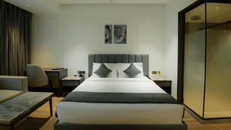 A king size bed flanked by nightstands, a study desk with a chair, and a glass partition at Awesome Palace, Guwahati Airport.