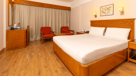 A room with a queen size bed, light wood floors, and a seating area with red armchairs near a window at Hotel Horizon, Trivandrum.