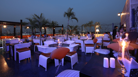 A rooftop dining area at dusk with white covered tables, chairs, burning candles, and lights reflecting on the floor at The Lindsay, Kolkata.
