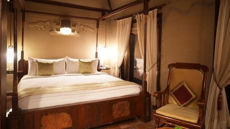 Dimly lit Zamindar Suite in Vedic Village Spa Resort showcasing a neatly made four-poster king size bed, wooden lounge chair, and high ceilings.