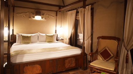 Dimly lit Zamindar Suite in Vedic Village Spa Resort showcasing a neatly made four-poster king size bed, wooden lounge chair, and high ceilings.