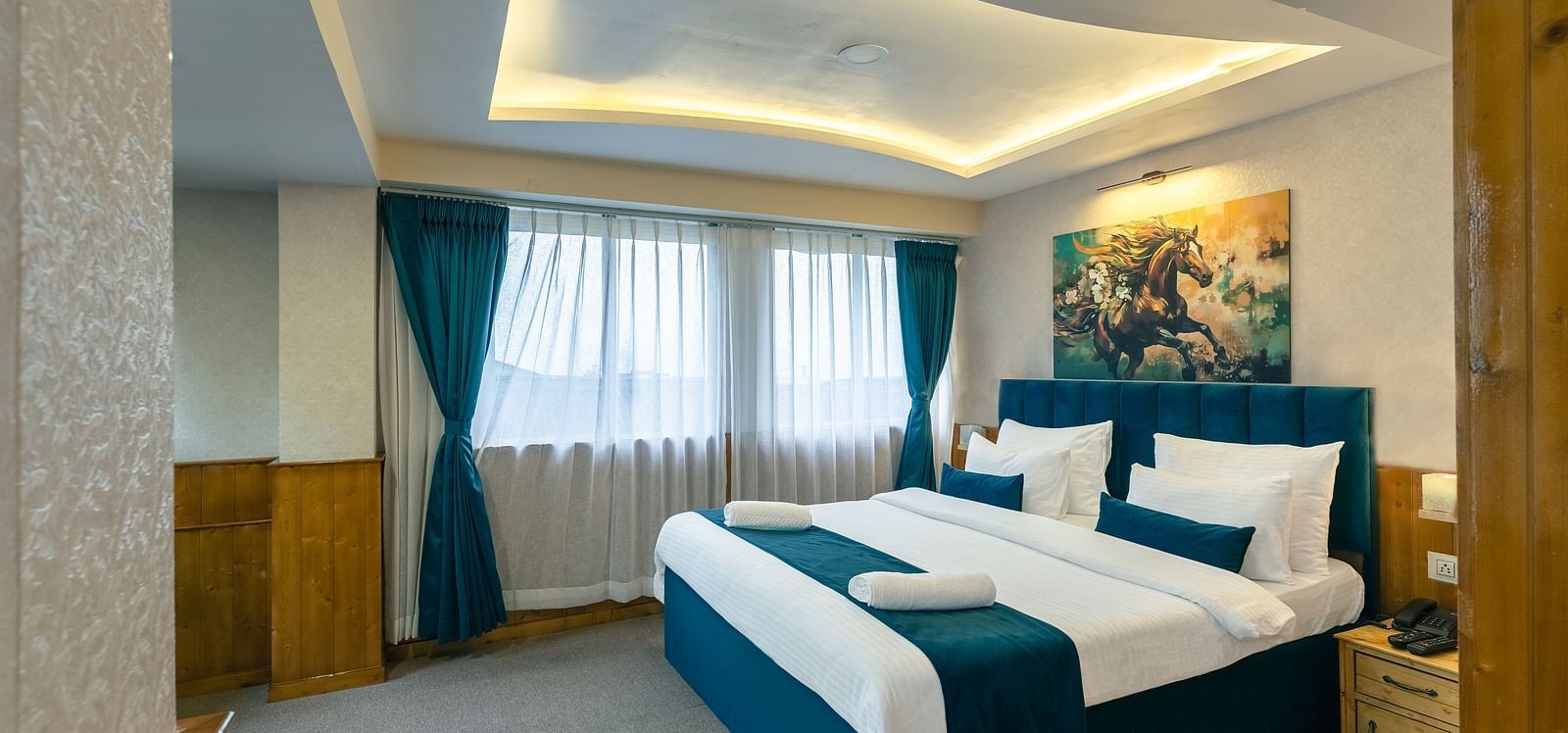 Deluxe Room with a king size bed, inviting ambiance, wooden bedside table, and a large window with curtains drawn at Avianna Seven Seas, Darjeeling.