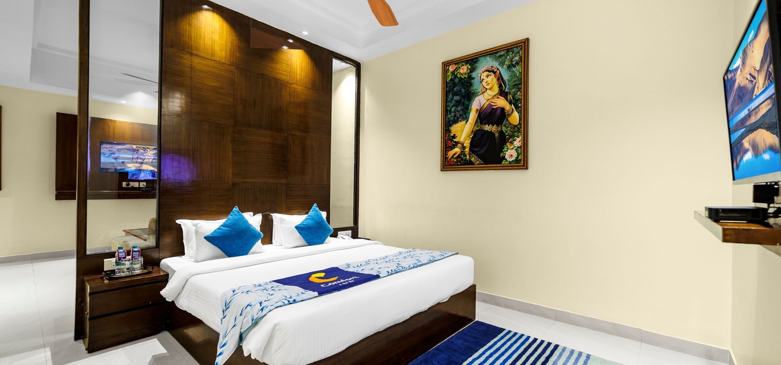 Bedroom at Inde Reva Resorts Rishhikesh with a double bed, blue cushions, ceiling fan, TV, and framed artwork on the wall.