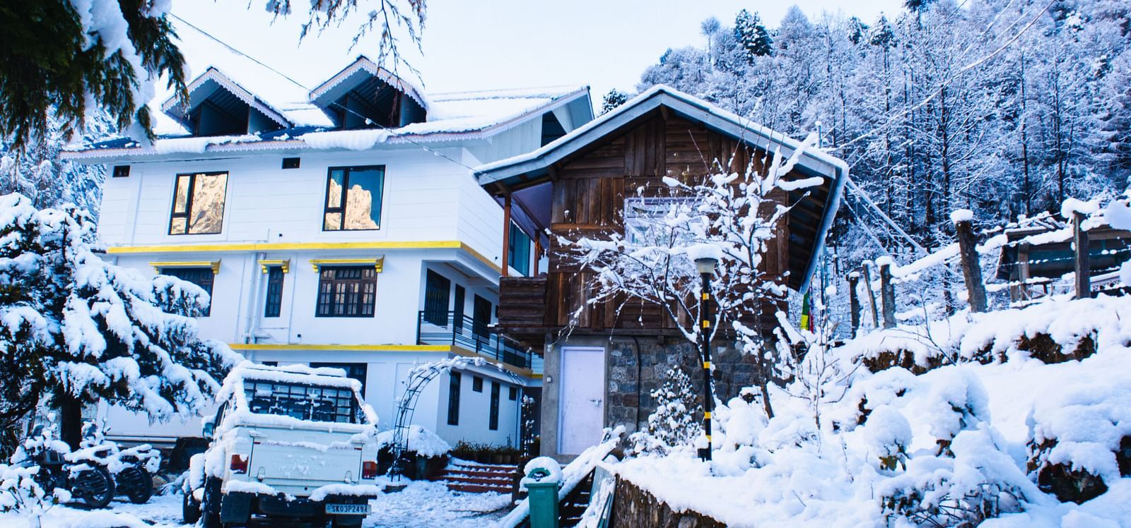 Snow-covered  Mystique Valley Resort nestled in a Himalayan forest, with a parked SUV, rustic stone path, and surrounding trees blanketed in white.