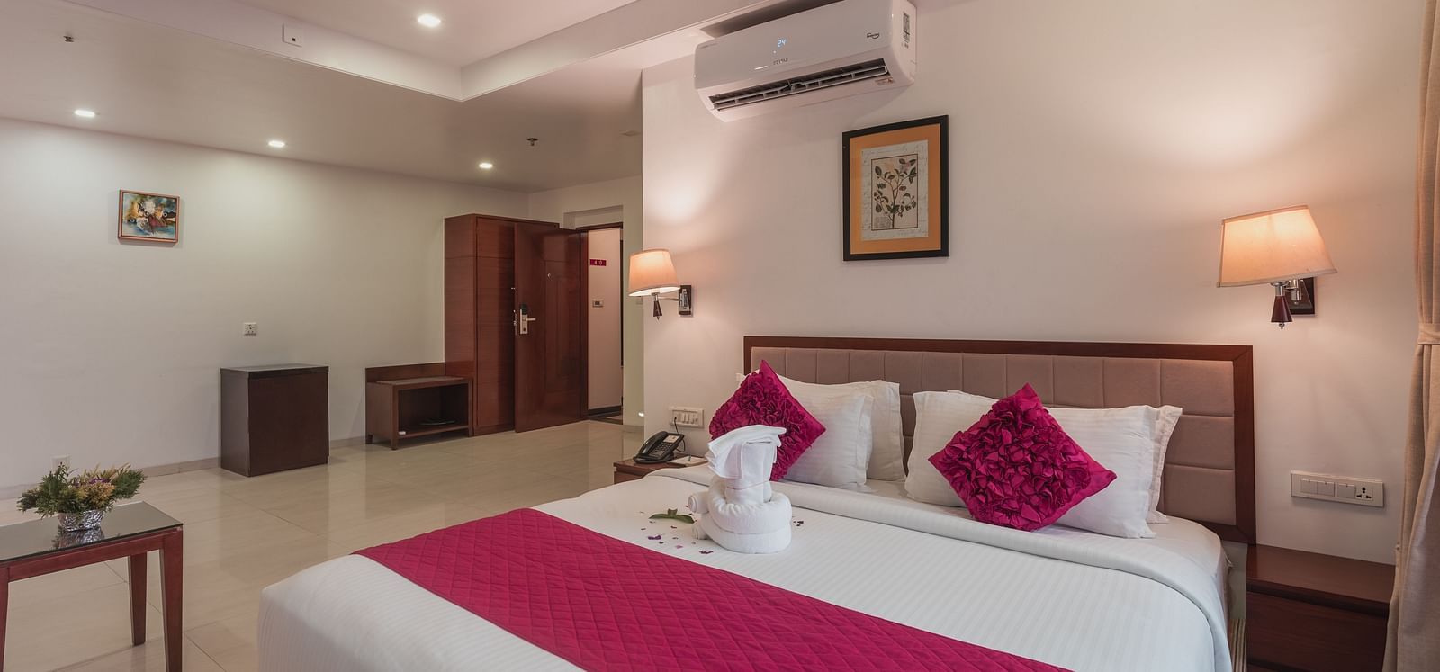 A hotel room at Diamond Palace Lords Plaza with a double bed accented with pink, a seating area, and wooden furniture.