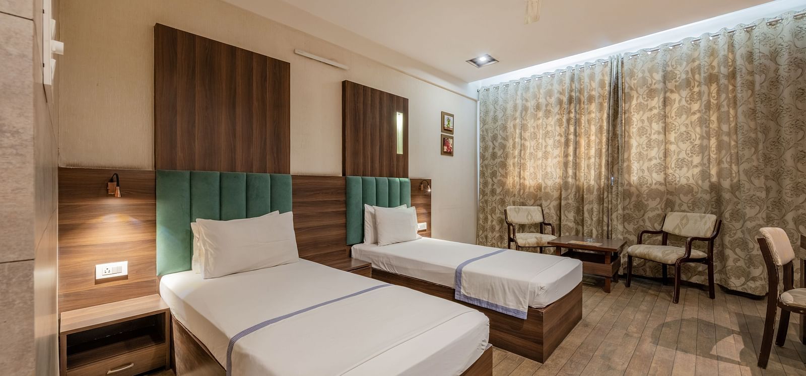 Twin beds with upholstered headboards and bedside tables beside a seating area for two and drawn curtains in the Maharaja Deluxe Rooms of Hotel Highway King, Behror.